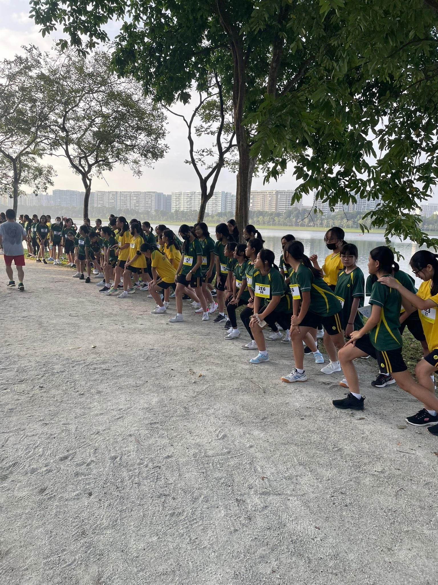 A large group of students in green and yellow uniforms crouch at a race starting line beside a lake with trees.
